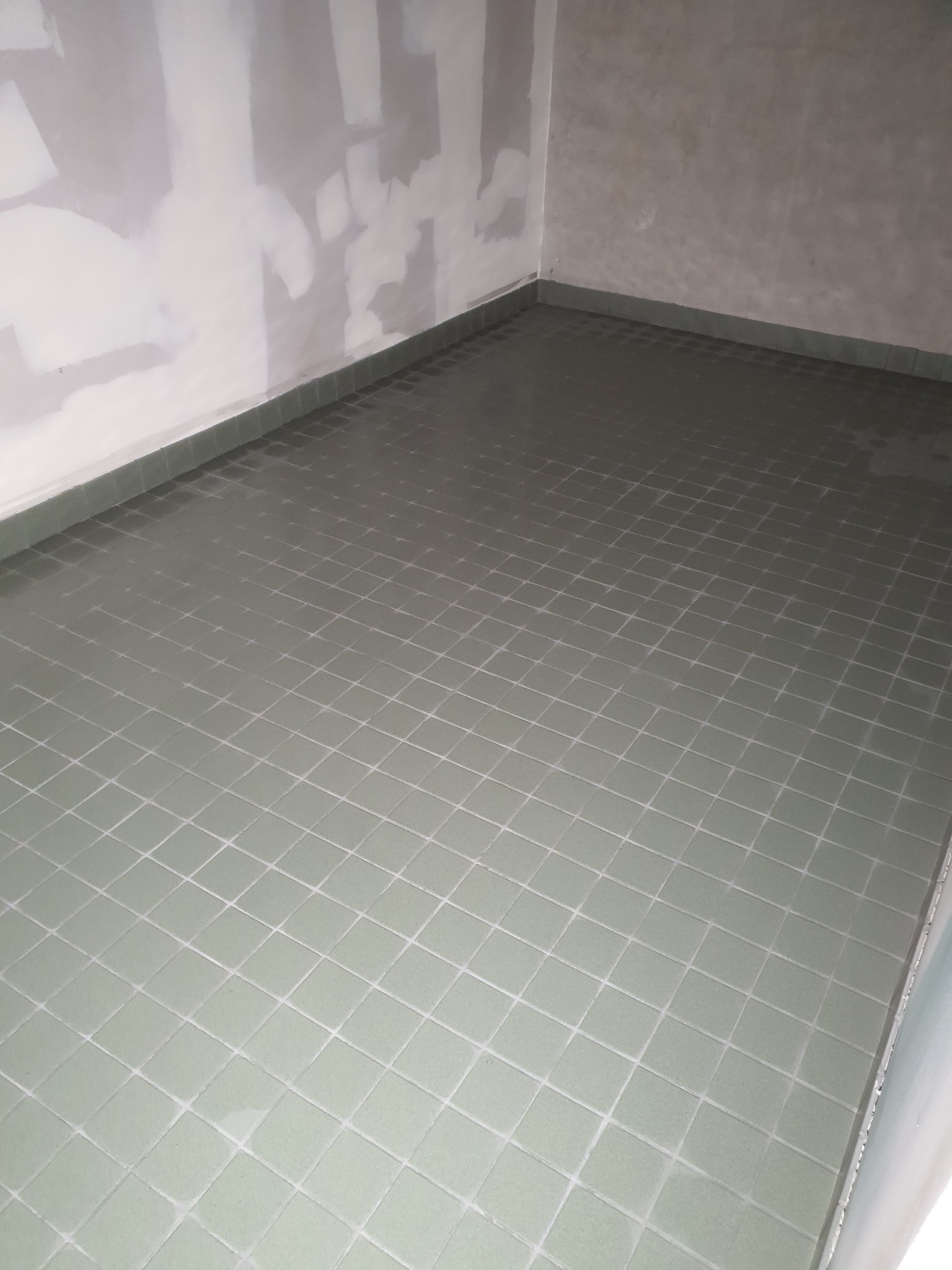 Świeżo położona, zielonkawa mozaika podłogowa w narożniku pomieszczenia, z widocznymi fugami i surowymi, częściowo wykończonymi ścianami.