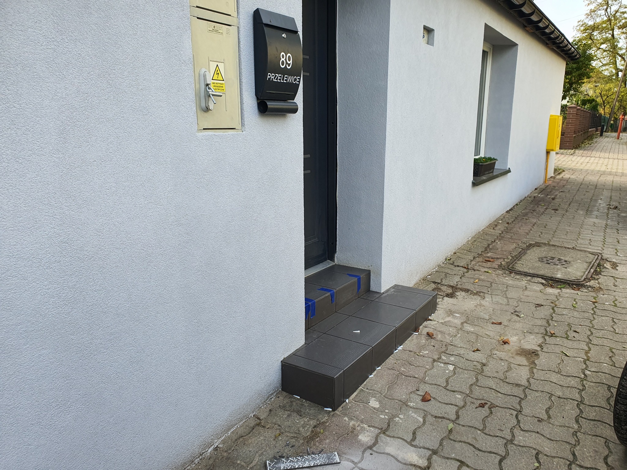 Nowe, ciemnoszare płytki na schodach wejściowych do domu, zabezpieczone niebieską taśmą, widoczne białe krzyżyki dystansowe, skrzynka na listy i tabliczka z numerem 89 Przelewice.