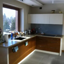 Nowoczesna kuchnia z drewnianymi szafkami, jasnym blatem i dużym oknem z widokiem na zieleń. Na blacie kwiaty i akcesoria kuchenne. Minimalistyczny design.