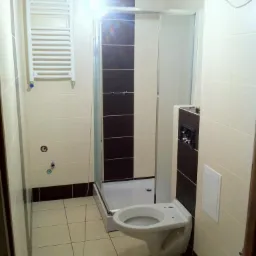 Wykończona łazienka z kabiną prysznicową i podwieszaną toaletą. Kremowe i brązowe płytki. Grzejnik drabinkowy na ścianie. Oświetlenie górne.