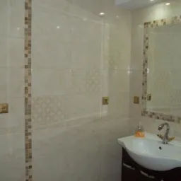 Jasna łazienka z beżowymi płytkami zdobionymi mozaiką, umywalka z szafką i lustro w ramie z mozaiki. Widoczne gniazdka elektryczne w złotych ramkach.