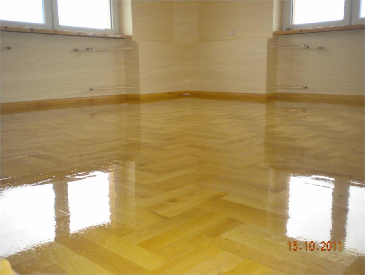 Wypolerowany, błyszczący parkiet ułożony w jodełkę w pustym pokoju z jasnymi ścianami i widocznymi rurami grzewczymi.