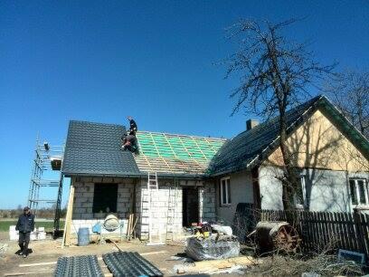 Budowa domu: Ekipa montuje pokrycie dachowe z blachy na nowym dachu, widoczne rusztowania, zielona membrana i przygotowane elementy pokrycia na ziemi.