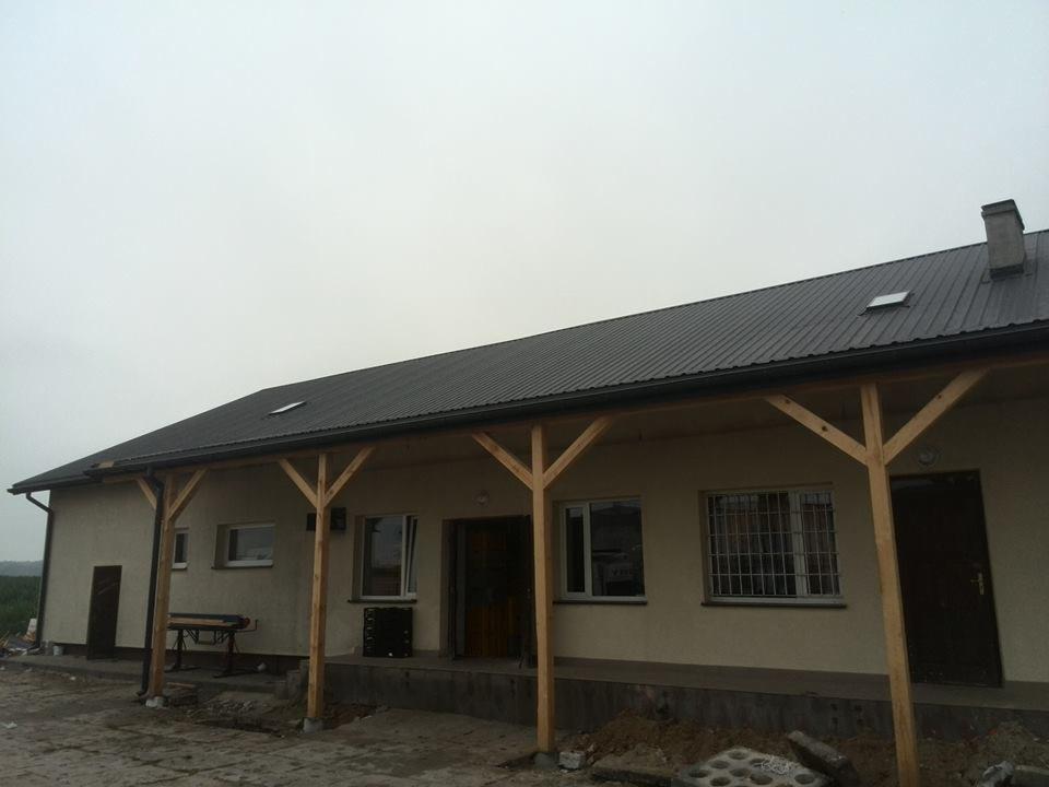 Budynek z ciemnoszarym dachem z blachy trapezowej, drewnianą konstrukcją zadaszenia i beżową elewacją. Widoczny komin, okna z kratami i ziemia wokół budynku.