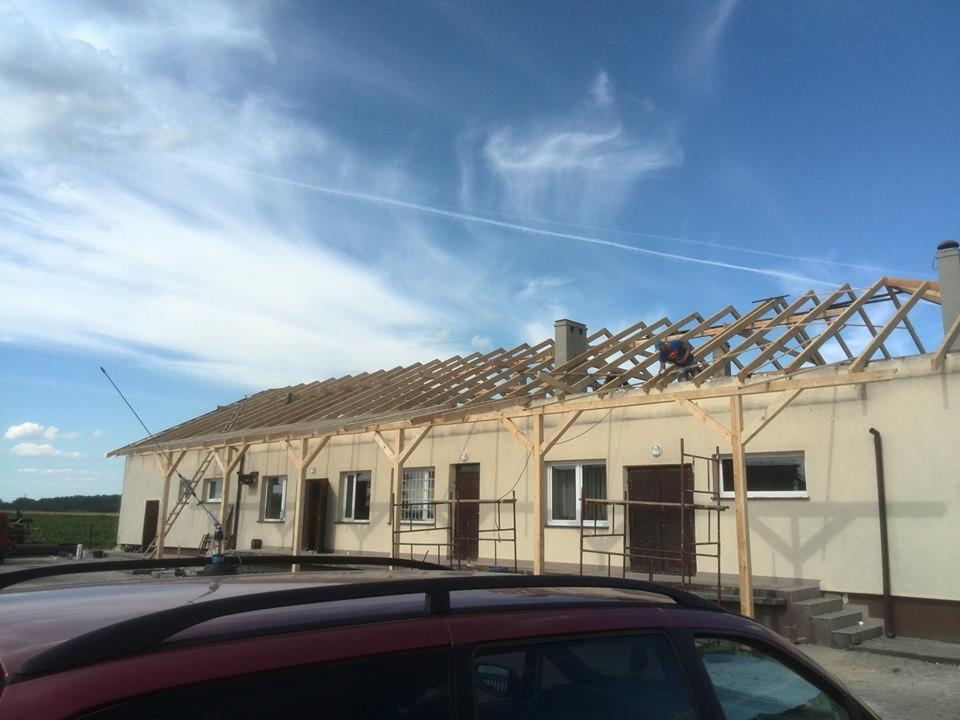 Budynek z drewnianą konstrukcją dachu w trakcie budowy, widoczny pracownik na dachu, fragment dachu samochodu na pierwszym planie, błękitne niebo w tle.