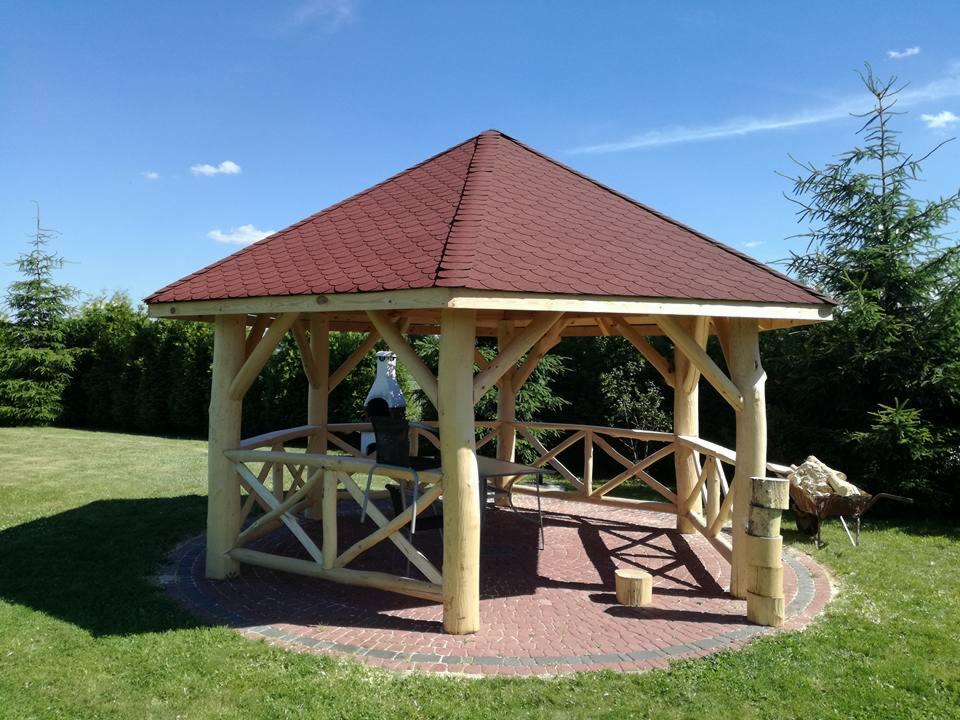 Drewniana altana ogrodowa z czerwonym dachem z gontu bitumicznego, z grillem wewnątrz i stosem pociętego drewna obok, na trawniku w słoneczny dzień.