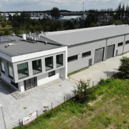 Nowoczesny budynek usługowy z częścią biurową i halą, widok z lotu ptaka, szara kostka brukowa, ogrodzenie z siatki, w tle zabudowania przemysłowe.