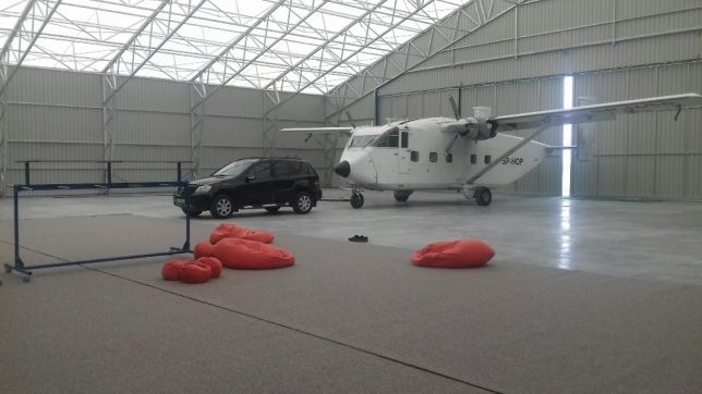 Wnętrze metalowej hali z widoczną konstrukcją dachu, w której stoi mały samolot, czarny SUV, kilka czerwonych puf i metalowy stelaż.