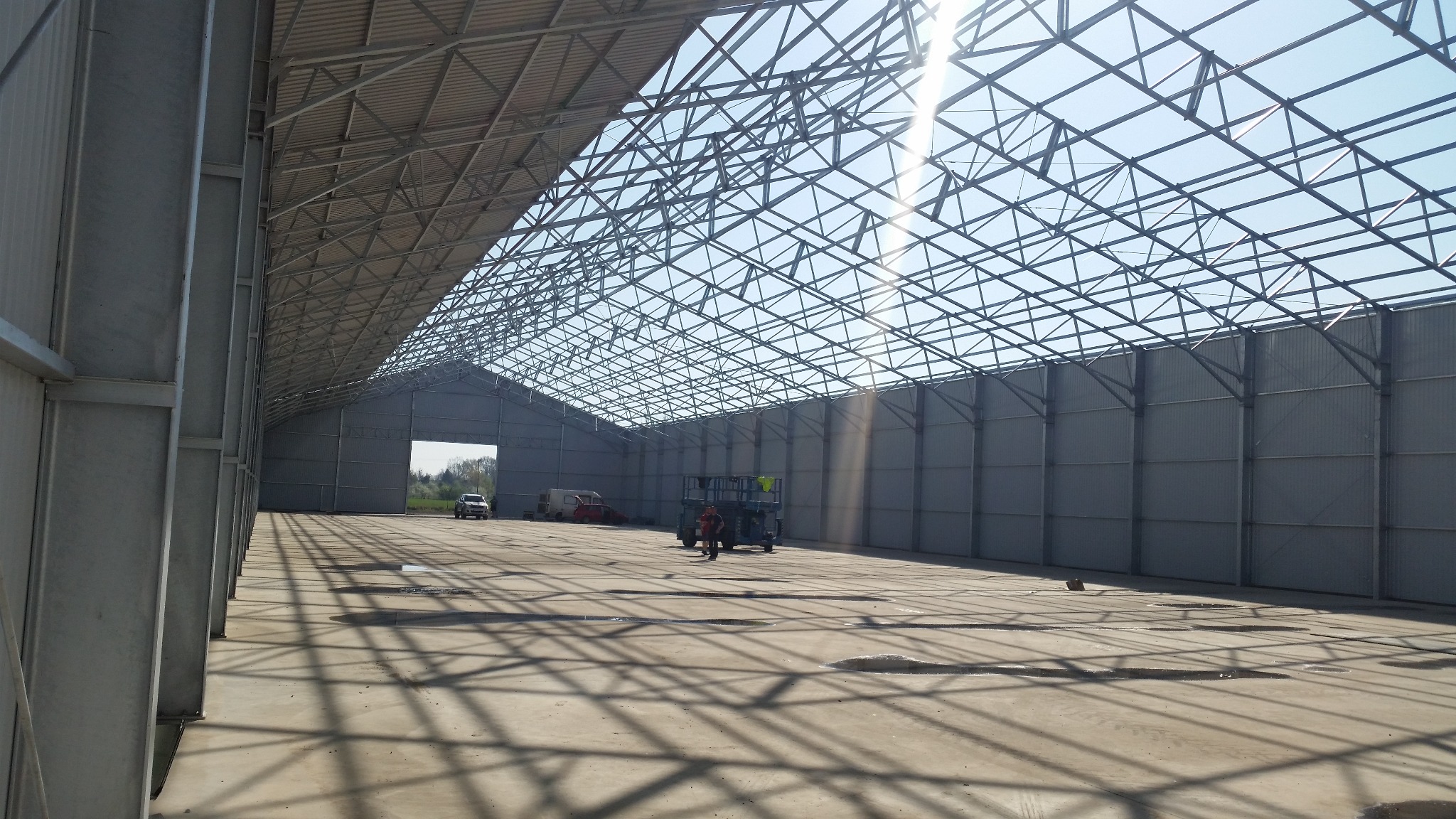 Wnętrze hali stalowej w trakcie budowy, widoczna konstrukcja dachu, ściany z blachy trapezowej, na posadzce cienie konstrukcji, w tle pojazdy i pracownik obsługujący podnośnik.