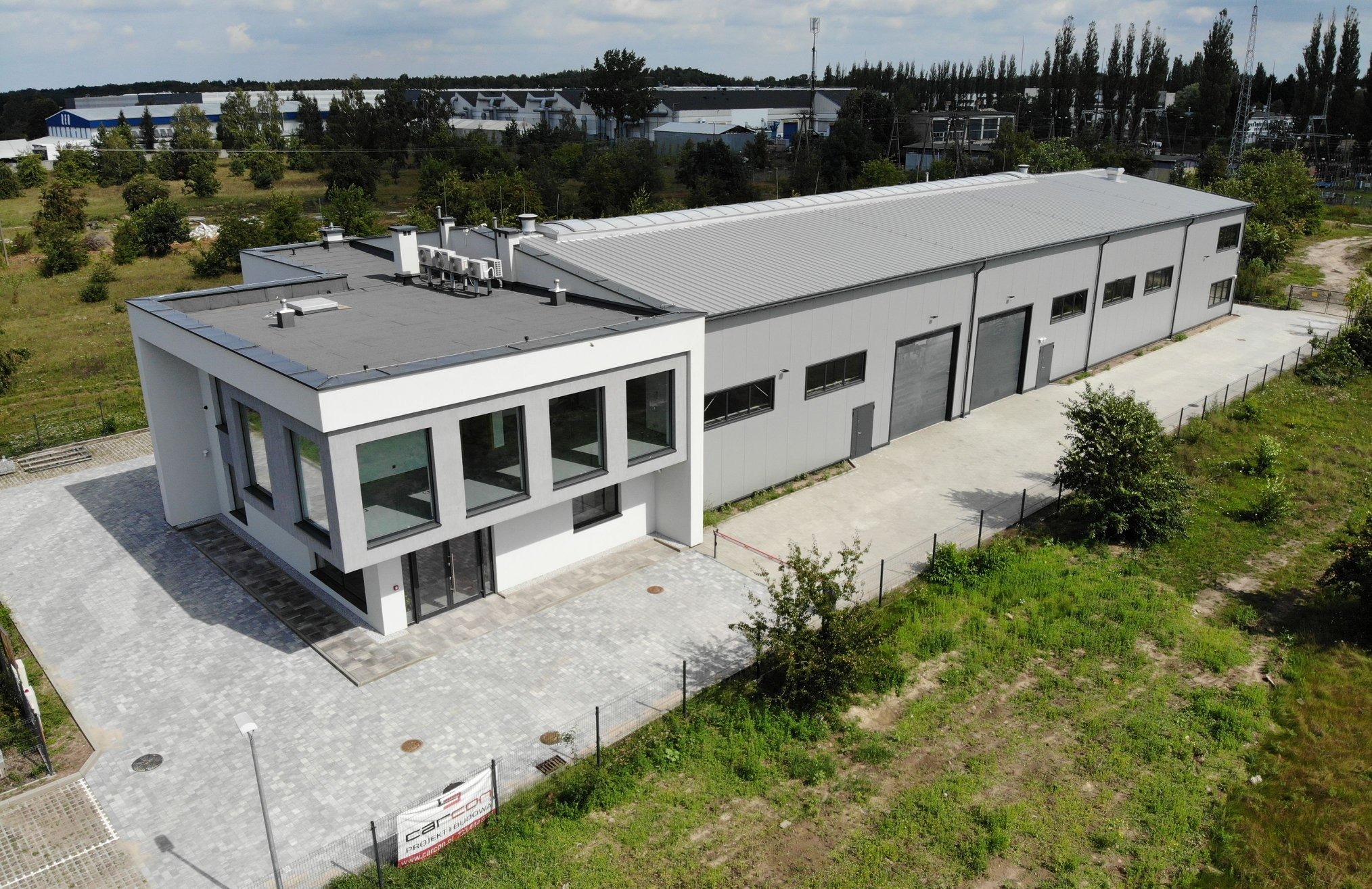 Nowoczesny budynek usługowy z częścią biurową i halą, widok z lotu ptaka, szara kostka brukowa, ogrodzenie z siatki, w tle zabudowania przemysłowe.