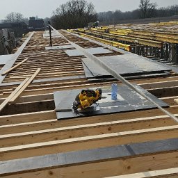 Developartus - Konstrukcja szalunkowa stropu z drewnianych belek, czarnych płyt i żółtego rusztowania. Na pierwszym planie piła tarczowa i butelka wody. W tle pracownik budowlany.