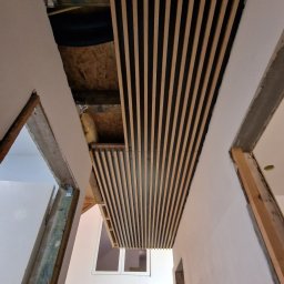 Developartus - Montaż dekoracyjnych paneli lamelowych na suficie w surowym wnętrzu. Widoczne elementy konstrukcyjne i okna w tle. Ujęcie z dołu, podkreślające perspektywę.