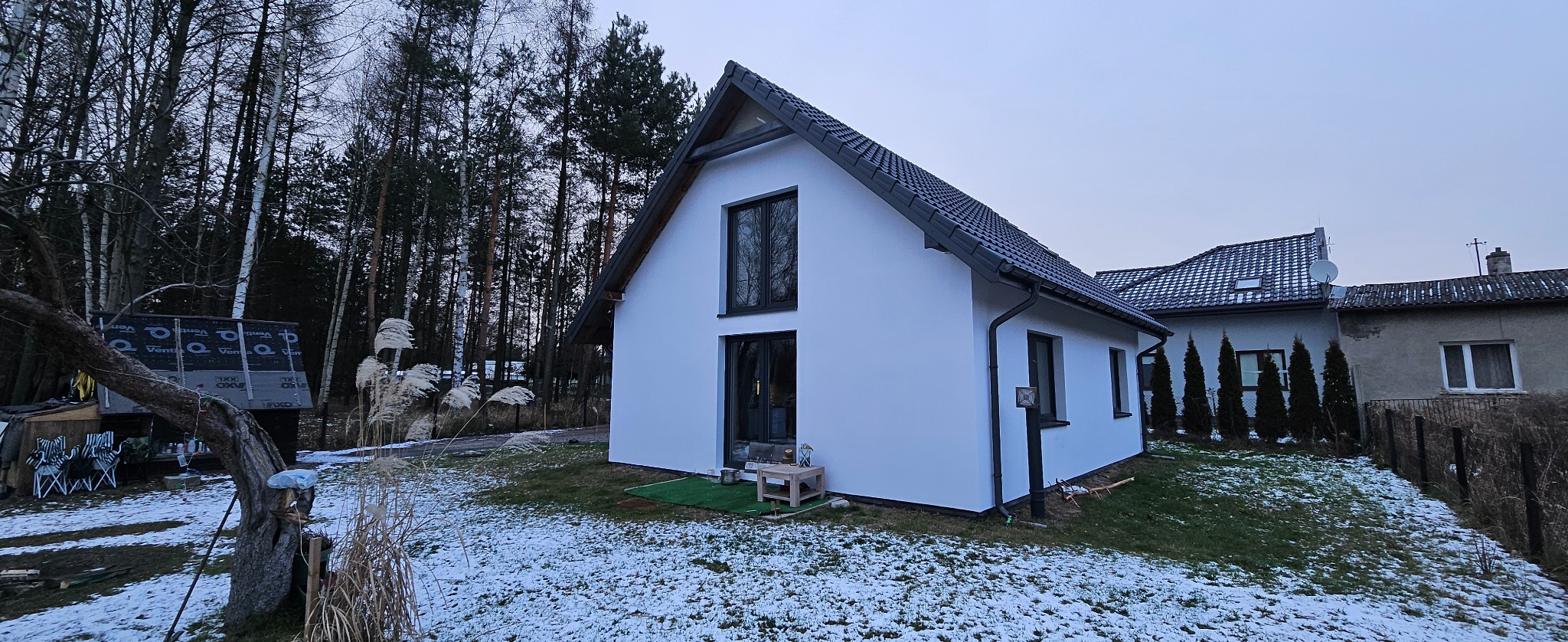 Nowoczesny, biały dom jednorodzinny z ciemną stolarką okienną i dachem na tle zimowego krajobrazu z drzewami. Widoczny fragment sąsiedniego budynku.