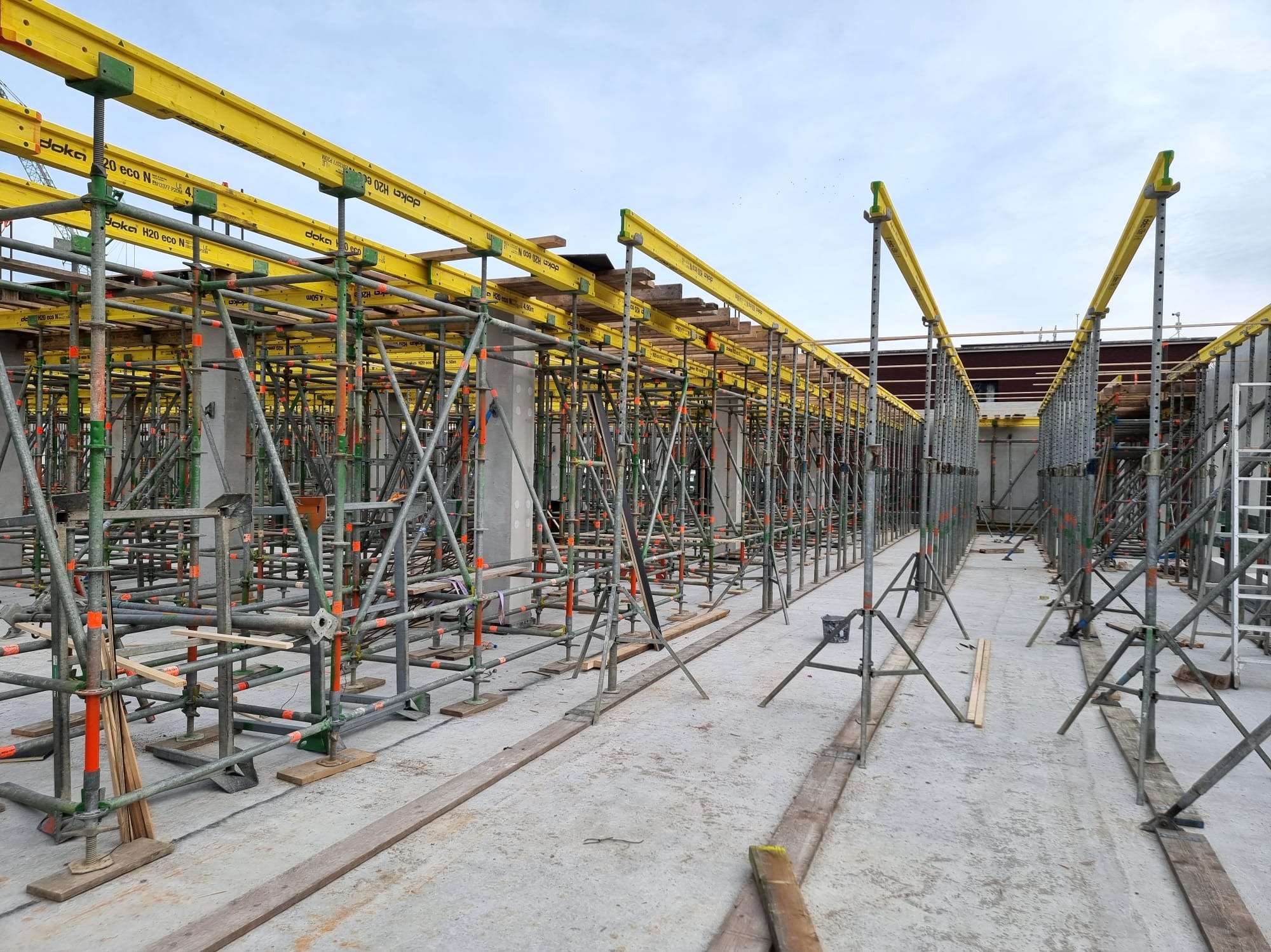 Szare rusztowania z żółtymi elementami Doka H20 eco N podtrzymujące konstrukcję stropu na budowie. Widok z dołu na konstrukcję, betonowa podłoga.