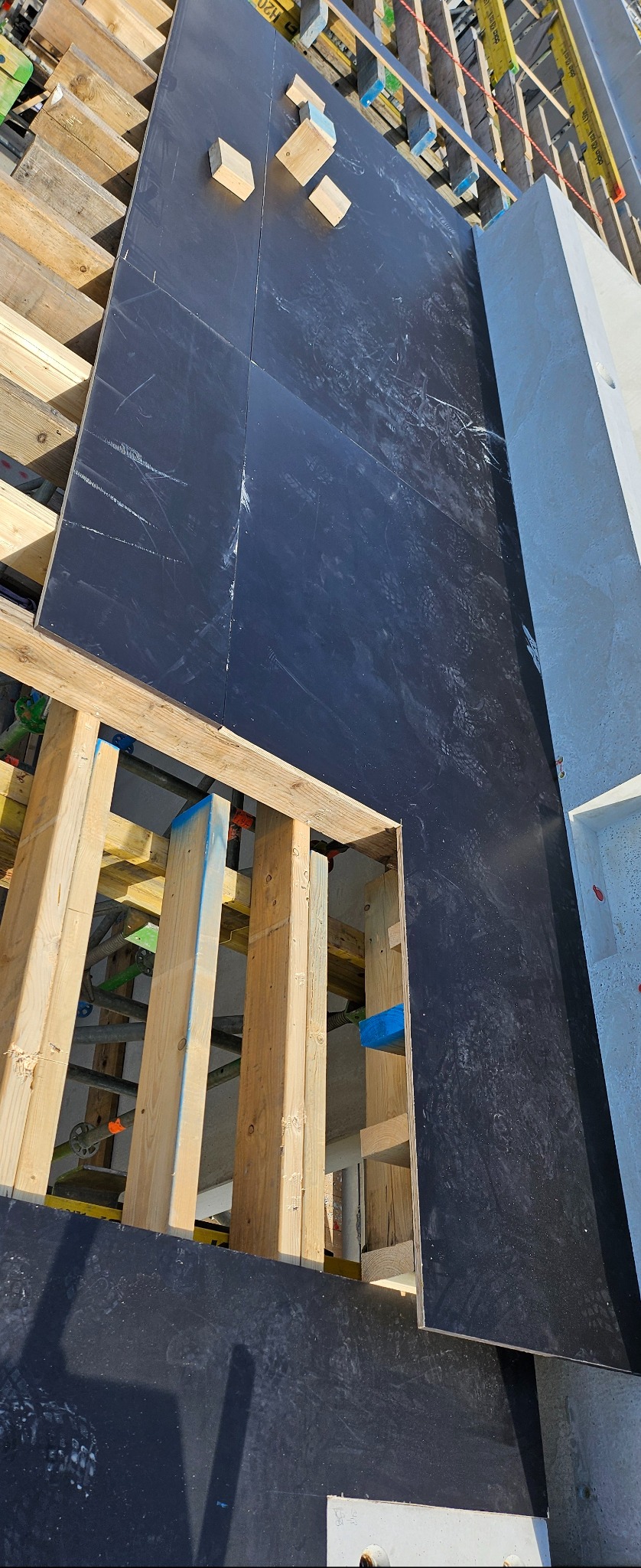 Szczegółowe ujęcie szalunku z desek i płyt na budowie, widoczne elementy konstrukcyjne i betonowy filar. Perspektywa z góry ukazuje etap prac budowlanych.