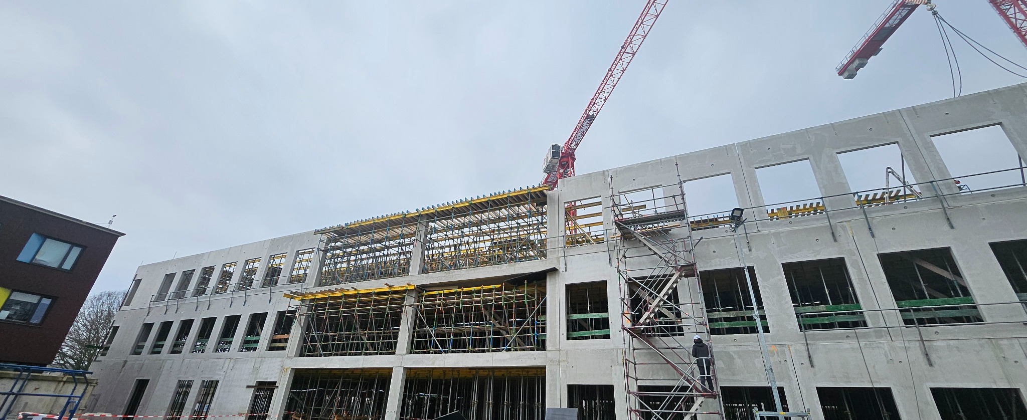 Budowa budynku żelbetowego z widocznymi szalunkami i rusztowaniami. Na pierwszym planie drabina rusztowaniowa z pracownikiem w kasku. W tle żurawie budowlane.