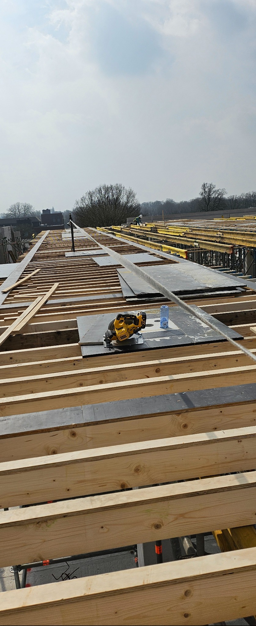 Konstrukcja szalunkowa stropu z drewnianych belek, czarnych płyt i żółtego rusztowania. Na pierwszym planie piła tarczowa i butelka wody. W tle pracownik budowlany.