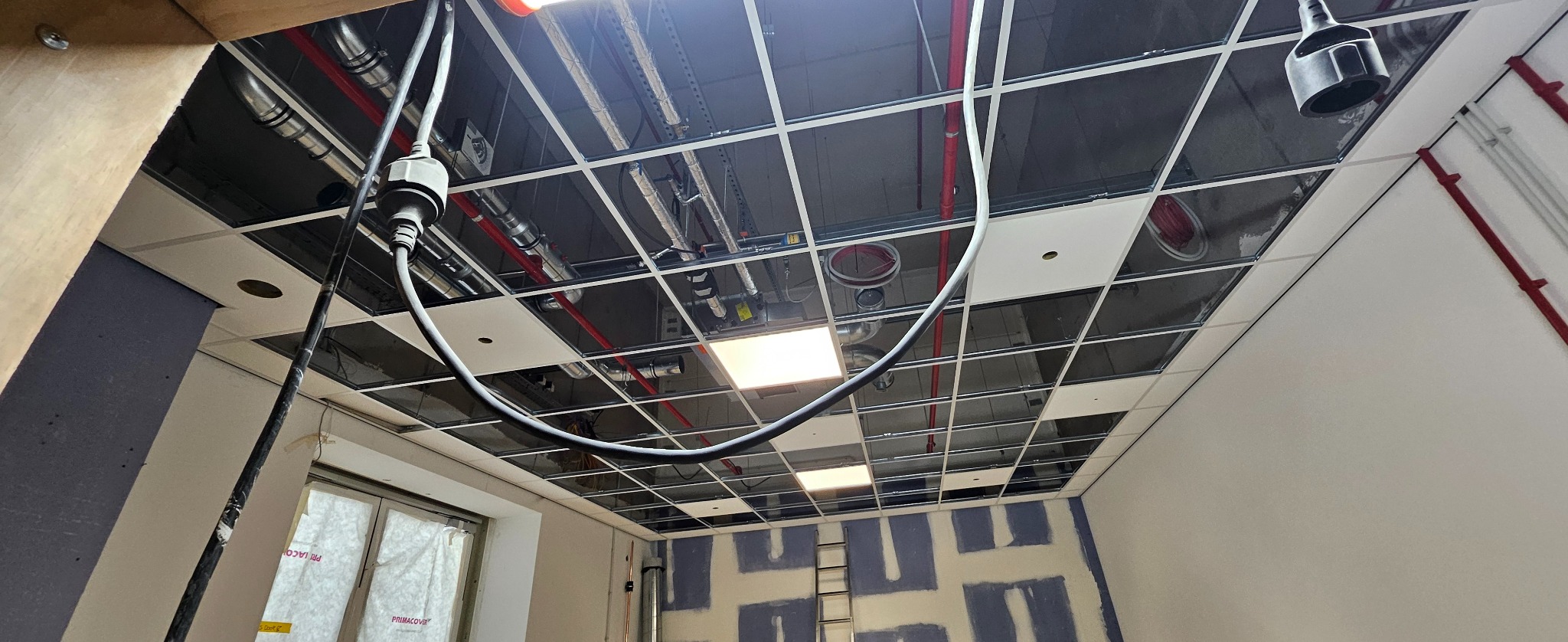 Montaż sufitu podwieszanego kasetonowego z widocznymi instalacjami wentylacyjnymi i elektrycznymi. Ściany przygotowane do malowania, widoczna drabina.