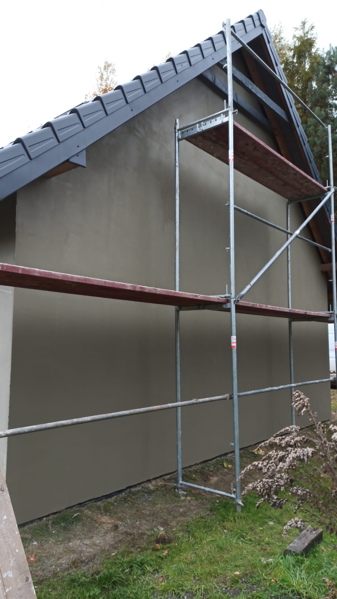 Świeżo otynkowana ściana domu jednorodzinnego z szarym dachem, widoczna z boku, z ustawionym rusztowaniem. Trawa i krzewy u podstawy ściany.