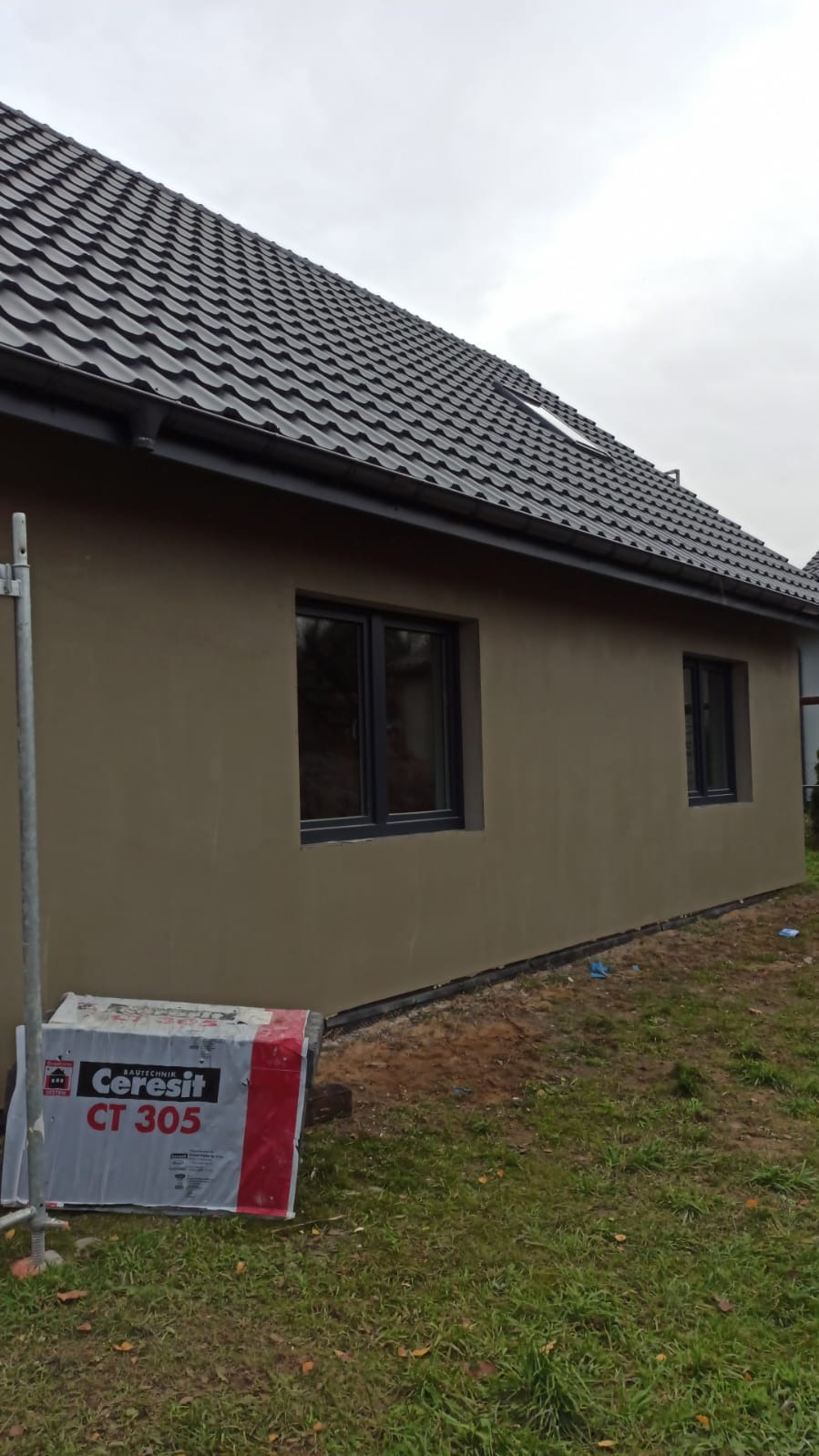 Ściana domu po nałożeniu tynku, widoczne okna z czarnymi ramami, dach z ciemnej dachówki i paleta z zaprawą Ceresit CT 305. Ujęcie z boku budynku.