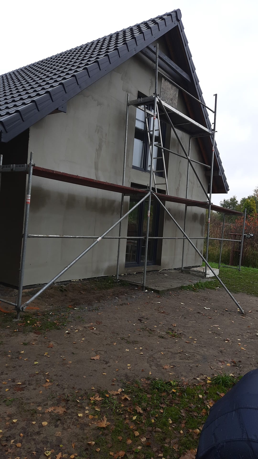 Dom jednorodzinny w trakcie prac elewacyjnych, widoczne rusztowania i świeżo nałożony tynk. Czarna dachówka kontrastuje z szarą elewacją. Jesienne liście na ziemi.