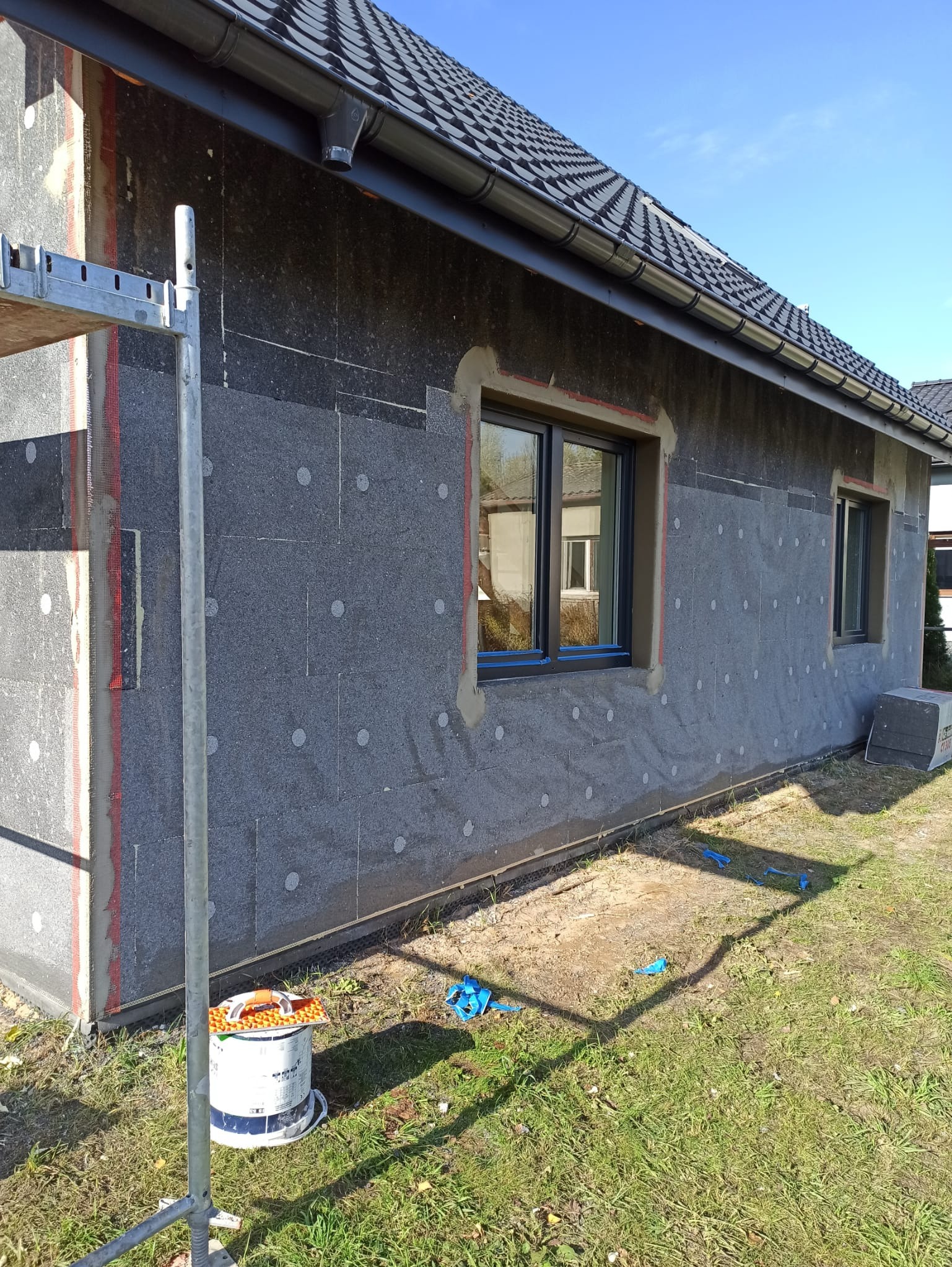 Ocieplanie elewacji domu jednorodzinnego z widocznymi płytami izolacyjnymi EPS, rusztowaniem i puszką farby. Prace w toku, widoczne okna i fragment dachu.