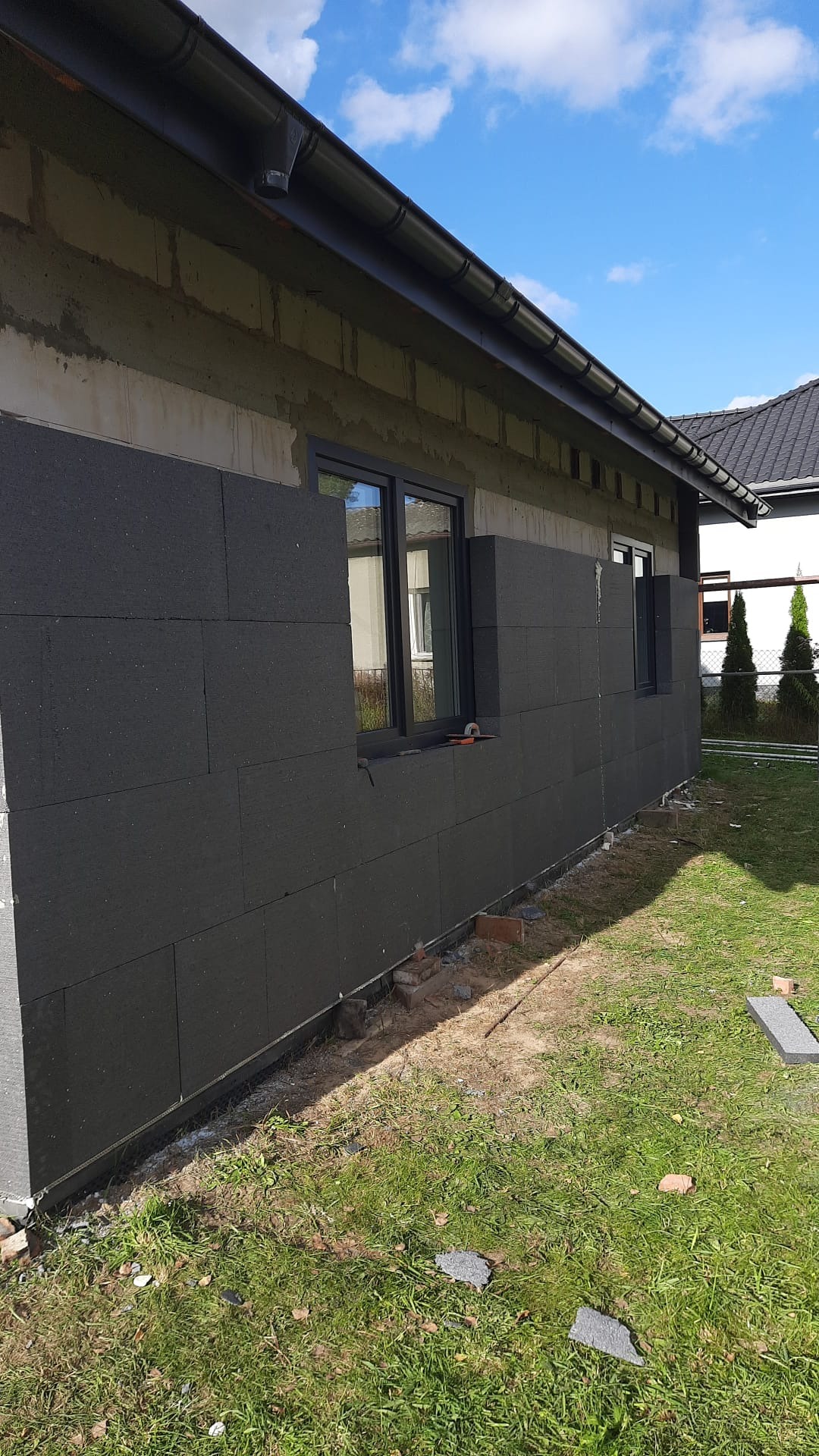 Ocieplanie elewacji domu jednorodzinnego grafitowym styropianem. Widoczne okna i fragment dachu. Prace w toku, na trawie leżą fragmenty materiału izolacyjnego.