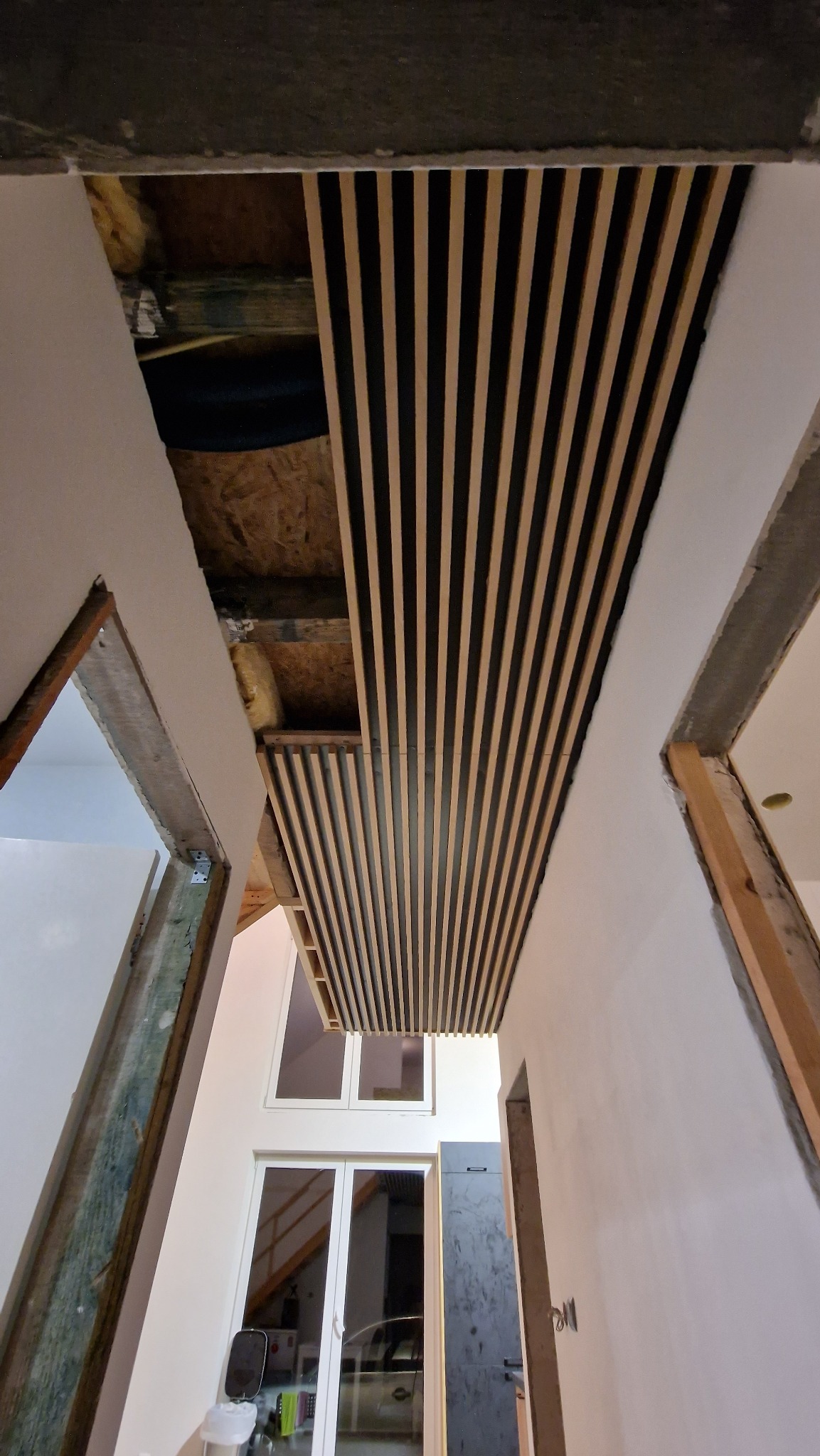 Montaż dekoracyjnych paneli lamelowych na suficie w surowym wnętrzu. Widoczne elementy konstrukcyjne i okna w tle. Ujęcie z dołu, podkreślające perspektywę.