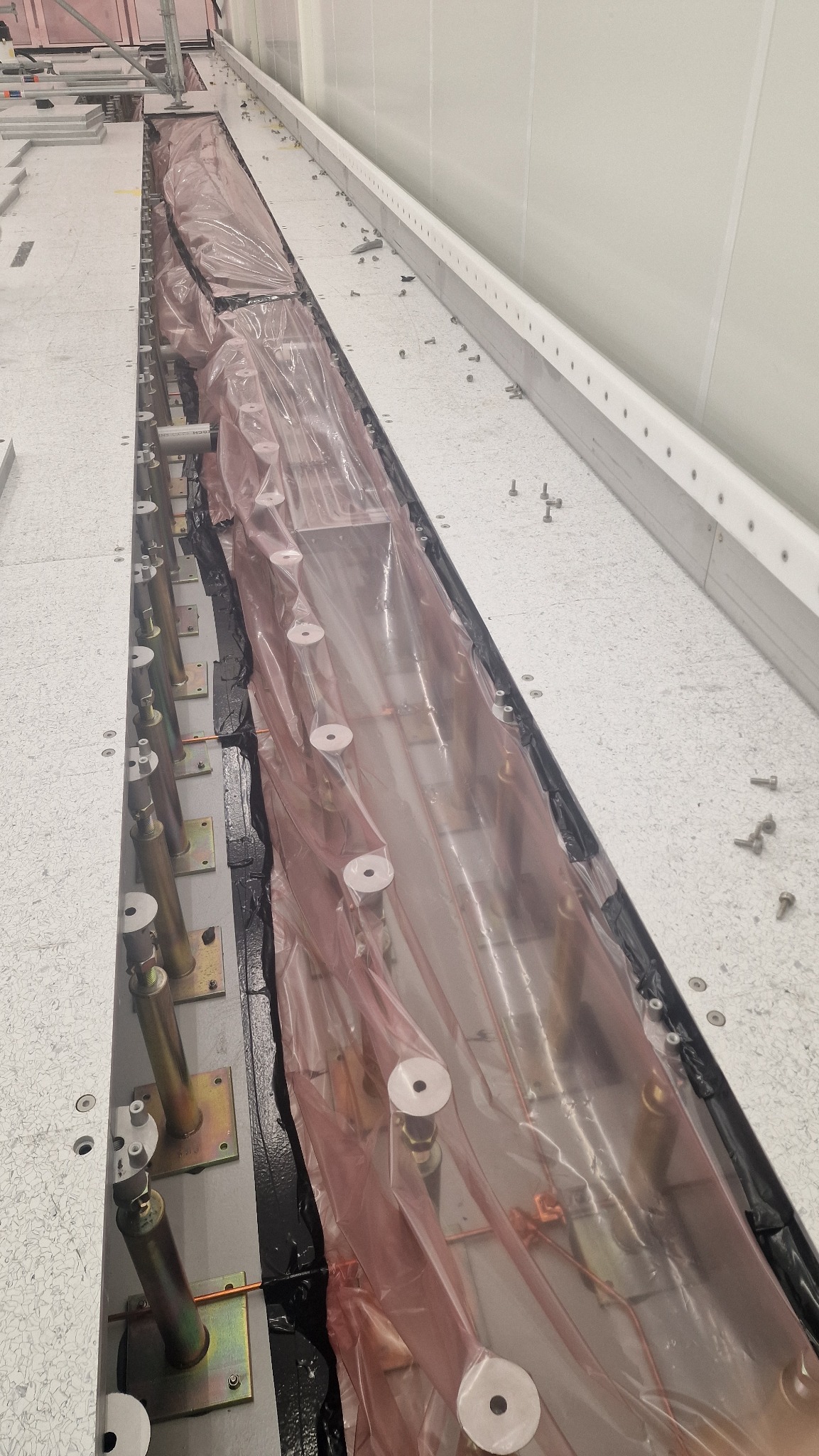 Wykopany rów z metalowymi wspornikami i rurami, zabezpieczony folią. Widoczne śruby i elementy montażowe na jasnej powierzchni. Prace instalacyjne wewnątrz budynku.