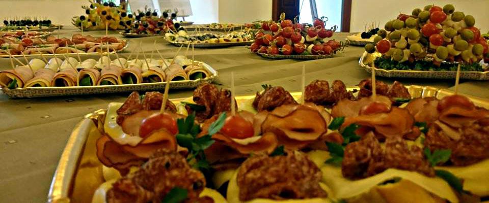 Stół z różnorodnymi przekąskami na srebrnych tacach: roladki z szynki z zielonym nadzieniem, koreczki z salami i pomidorkami koktajlowymi, piramida z oliwek i pomidorów, kompozycje owocowe.