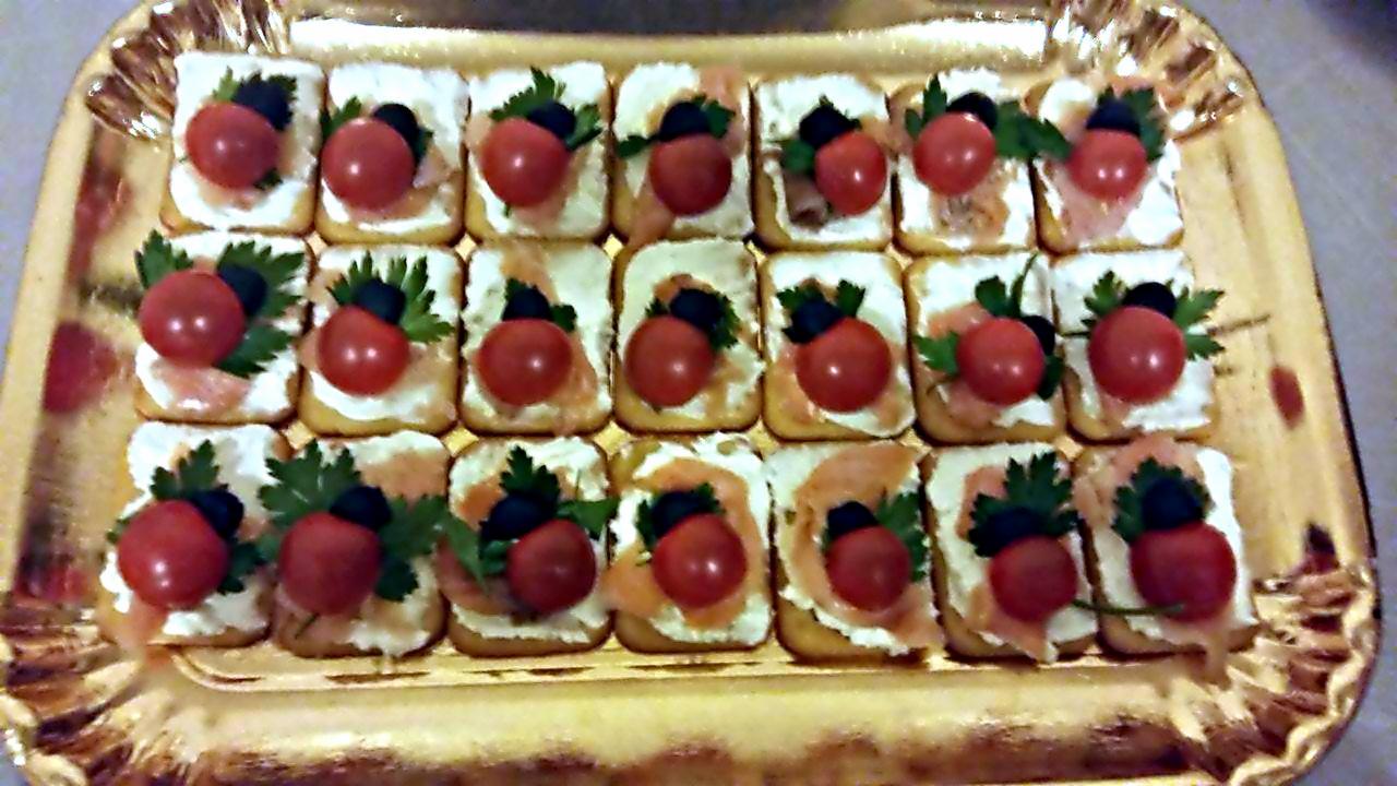 Kwadratowe kanapeczki na złotym talerzu: kremowy serek, łosoś, pomidorek cherry, oliwka i natka pietruszki.