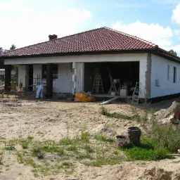 Budowa parterowego domu z garażem w trakcie ocieplania. Widoczny pracownik w kombinezonie oraz rusztowania i drabiny. Dach pokryty czerwoną dachówką.