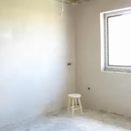 Wnętrze w trakcie prac wykończeniowych: świeżo otynkowane ściany w jasnym odcieniu, surowy sufit z drewnianymi belkami i plastikowe okno. Na podłodze stoi stołek.