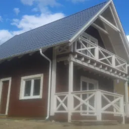 Dwukondygnacyjny dom z ciemnego drewna z białymi elementami wykończeniowymi i balkonem. Dach z szarej blachy, błękitne niebo w tle.