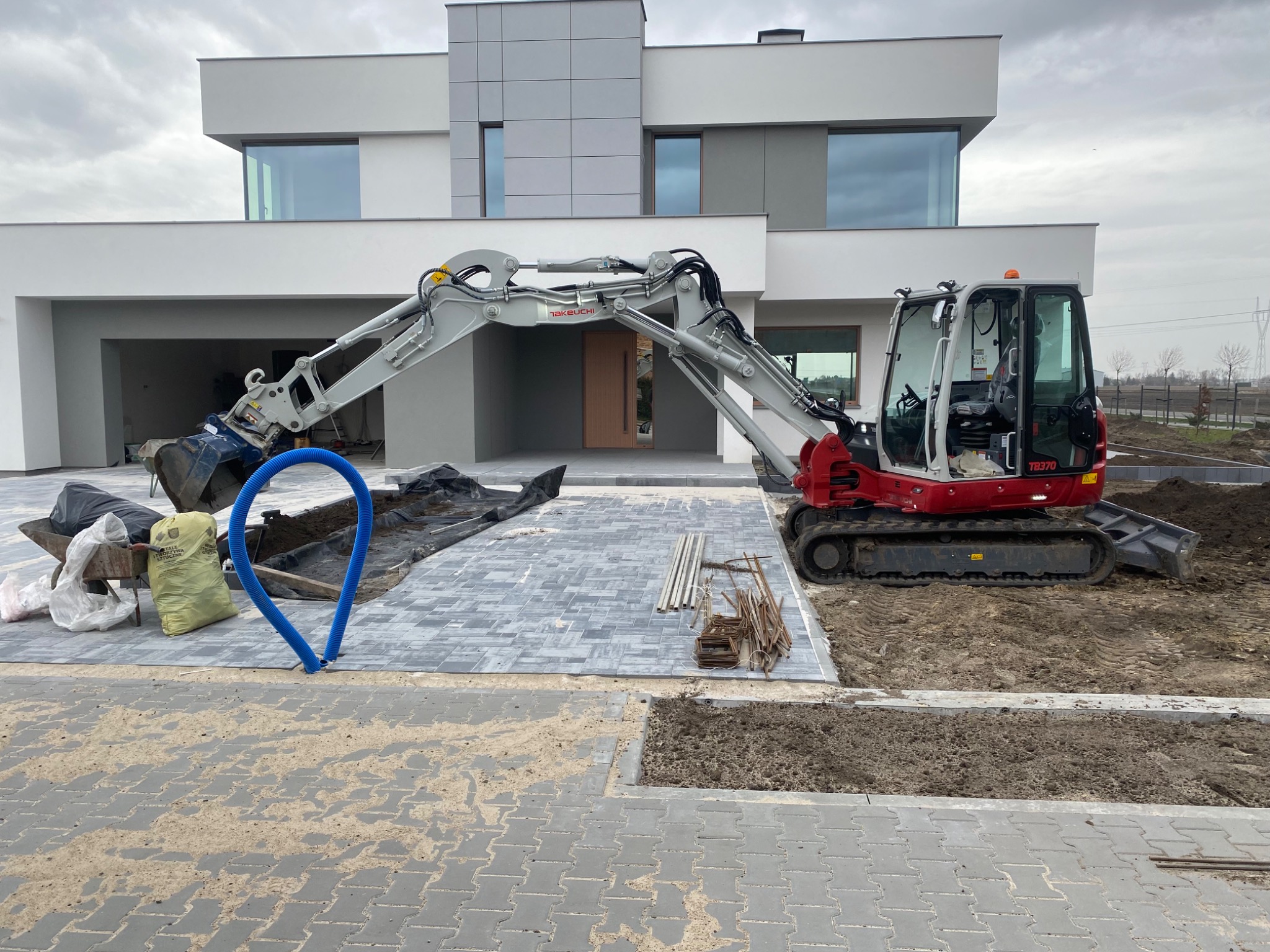 Czerwono-szara mini koparka Takeuchi TB370 na placu budowy przed nowoczesnym domem z szarymi płytami, przygotowująca teren pod podjazd z szarej kostki brukowej, widoczne worki, rury i narzędzia.