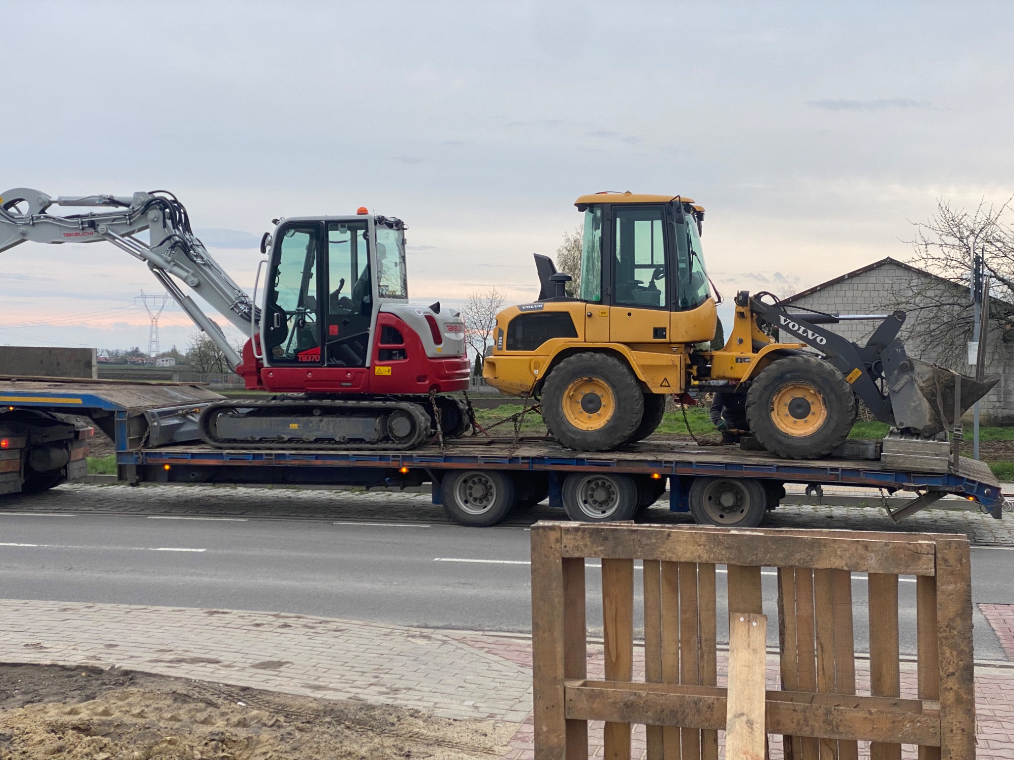 Transport niskopodwoziowy z koparką Takeuchi TB230 i ładowarką Volvo L30G na tle szarego nieba, widziane z poziomu chodnika wyłożonego kostką.