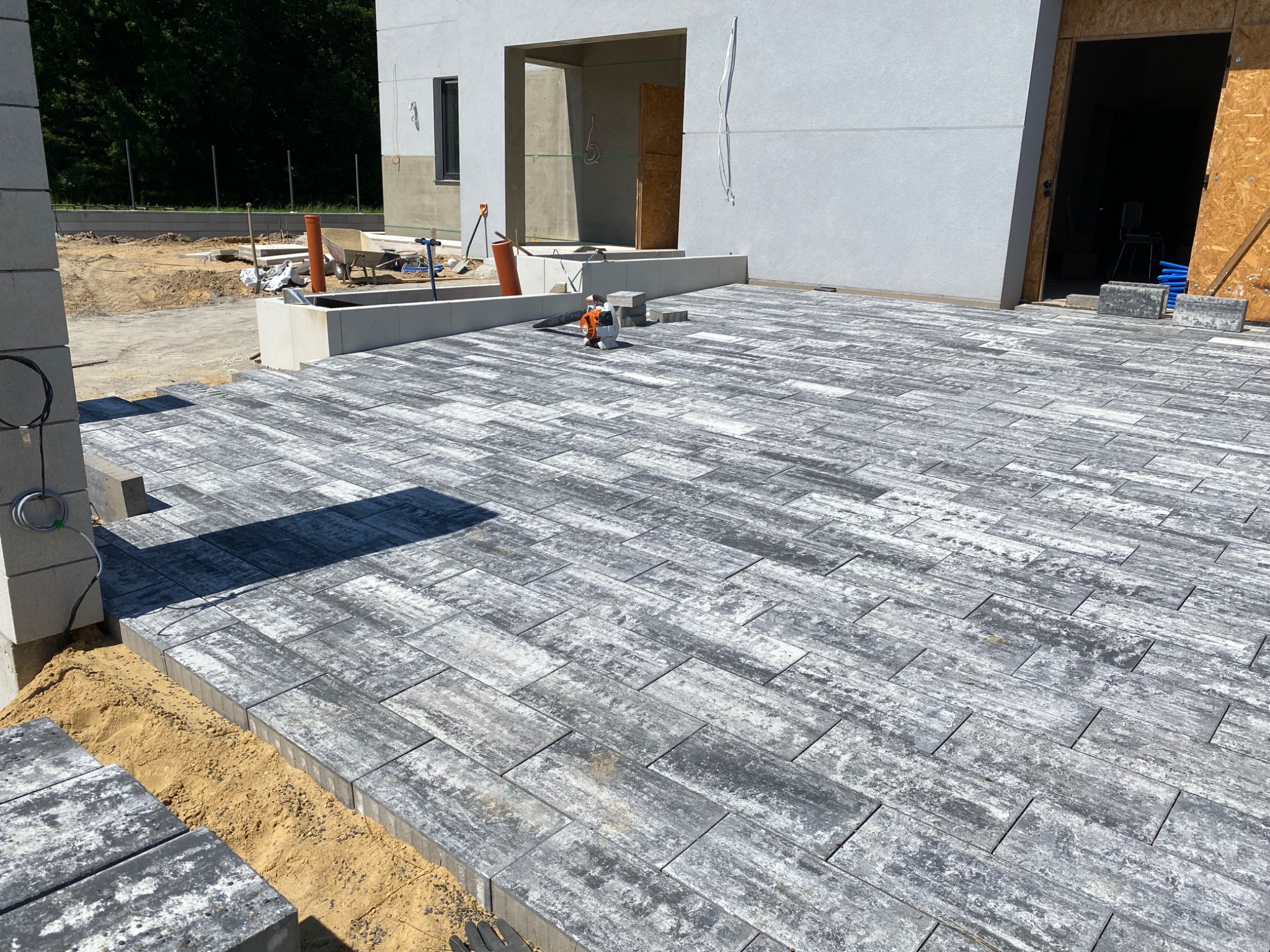 Ułożona szara kostka brukowa w jodełkę na posesji przed domem w trakcie budowy, widoczny piasek, narzędzia i niedokończone elementy.