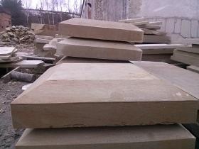 Stos bloków z piaskowca o zróżnicowanych kształtach i rozmiarach, ułożonych na zewnątrz, z widoczną stertą mniejszych elementów w tle i fragmentem budynku.