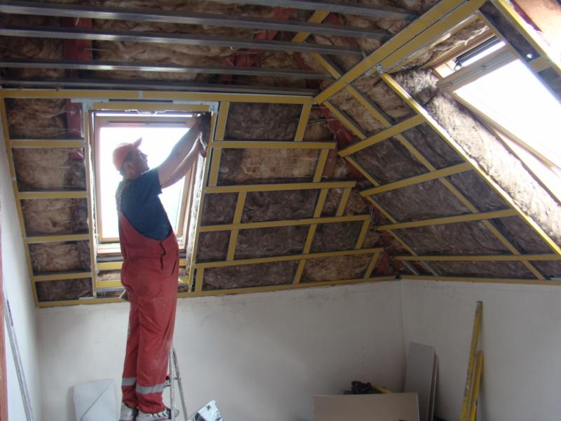 Pracownik w czerwonym kombinezonie montuje izolację z wełny mineralnej na poddaszu z oknem dachowym, widoczne metalowe profile konstrukcyjne i fragment drabiny.