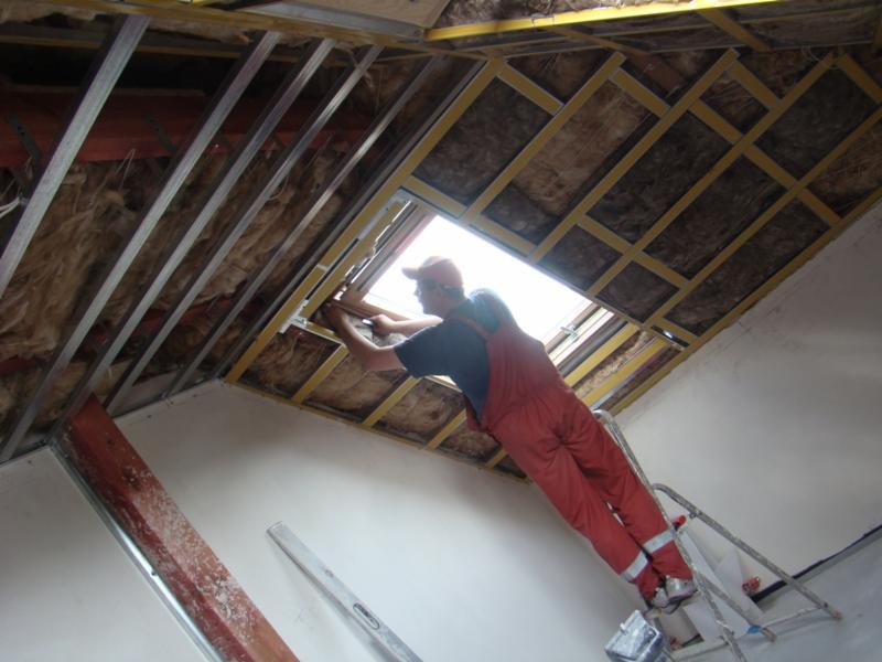 Pracownik w czerwonym kombinezonie na drabinie montuje okno dachowe w nowym budynku, widoczne metalowe profile i izolacja stropu.