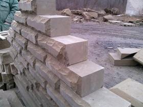 Fragment muru z piaskowca, widoczne surowe, obrobione bloki o nieregularnej fakturze, częściowo ułożone, z widocznym pracownikiem w tle po lewej stronie.