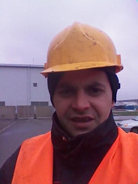 Pracownik w pomarańczowej kamizelce i żółtym kasku ochronnym na tle budynku przemysłowego i parkingu.