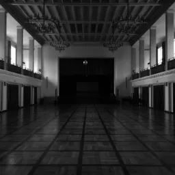 Czarno-białe zdjęcie przestronnej sali balowej z parkietem w szachownicę, kolumnami po bokach, balkonami na górze i żyrandolami zwisającymi z sufitu. Scena z ciemną kurtyną w tle.