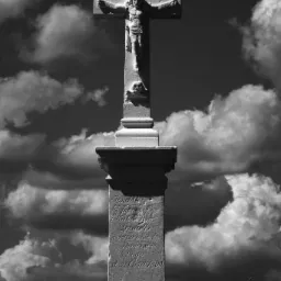 Czarno-białe zdjęcie kamiennego krzyża z Jezusem na tle pochmurnego nieba i pola pszenicy, z inskrypcjami na cokole.