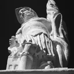 Czarno-białe zdjęcie z dołu przedstawiające kamienną rzeźbę postaci w szatach, z twarzą skierowaną ku górze i trzymającą w dłoniach coś w rodzaju zwoju lub księgi, na tle ciemnego nieba.