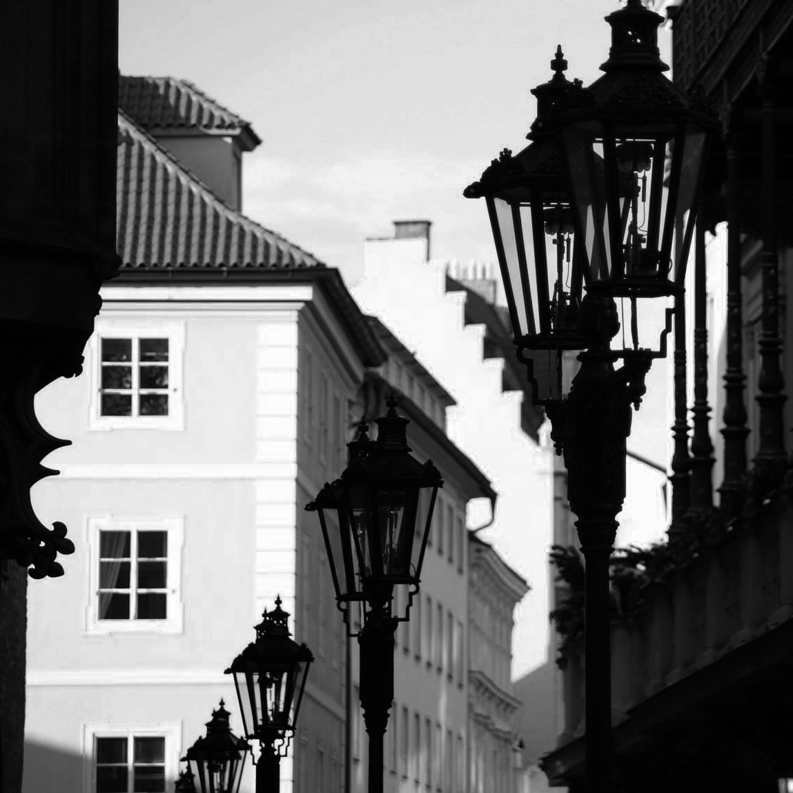 Czarno-biała fotografia ulicy z rzędem zabytkowych latarni gazowych, w tle budynki z oknami o regularnym układzie i schodkową architekturą dachu.