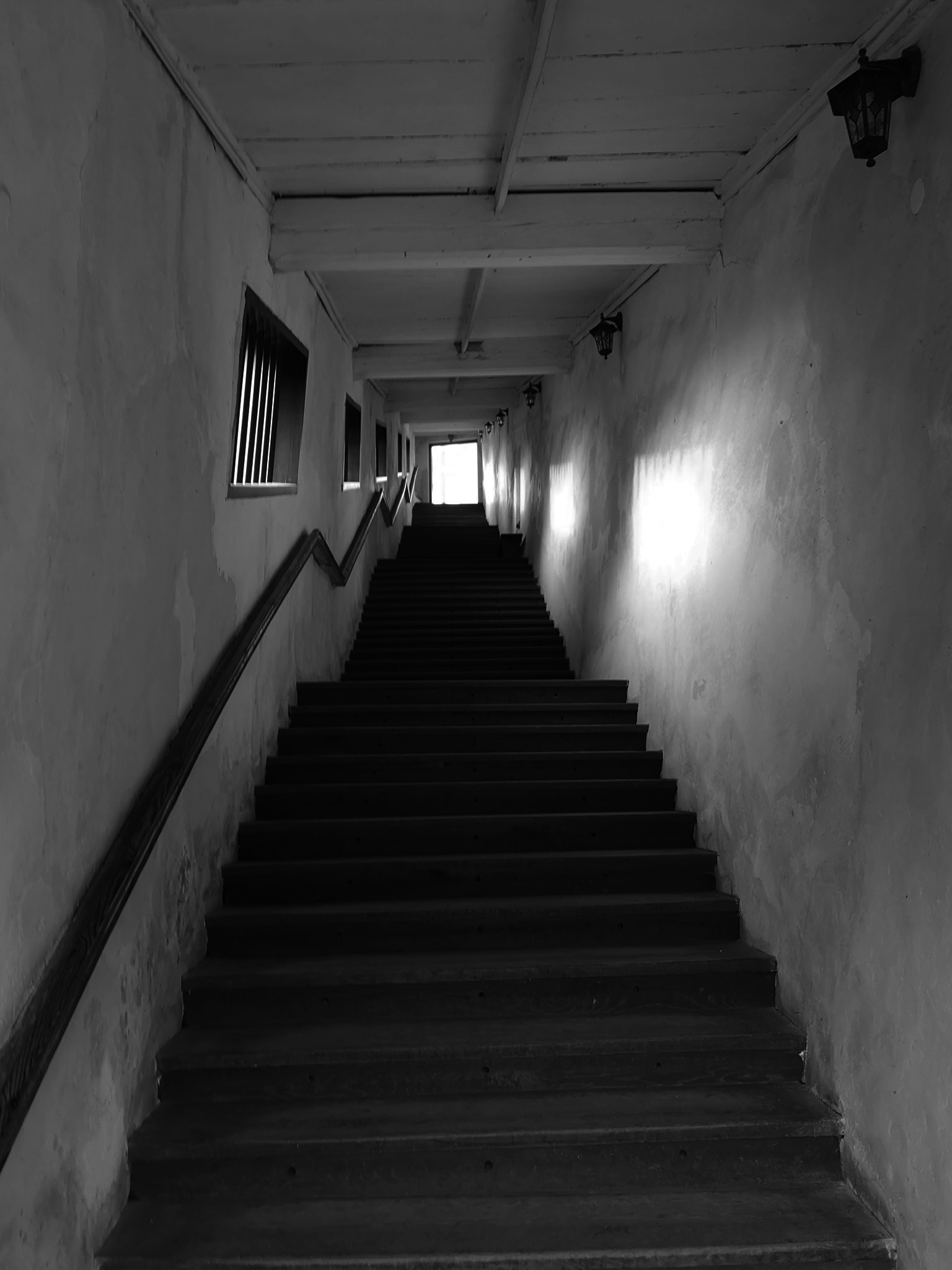 Wnętrze klatki schodowej w Sosnowcu z długimi, drewnianymi schodami prowadzącymi w górę, oświetlonymi przez okna i lampy ścienne w stylu retro. Ściany z wyraźną fakturą i drewniane belki stropowe.