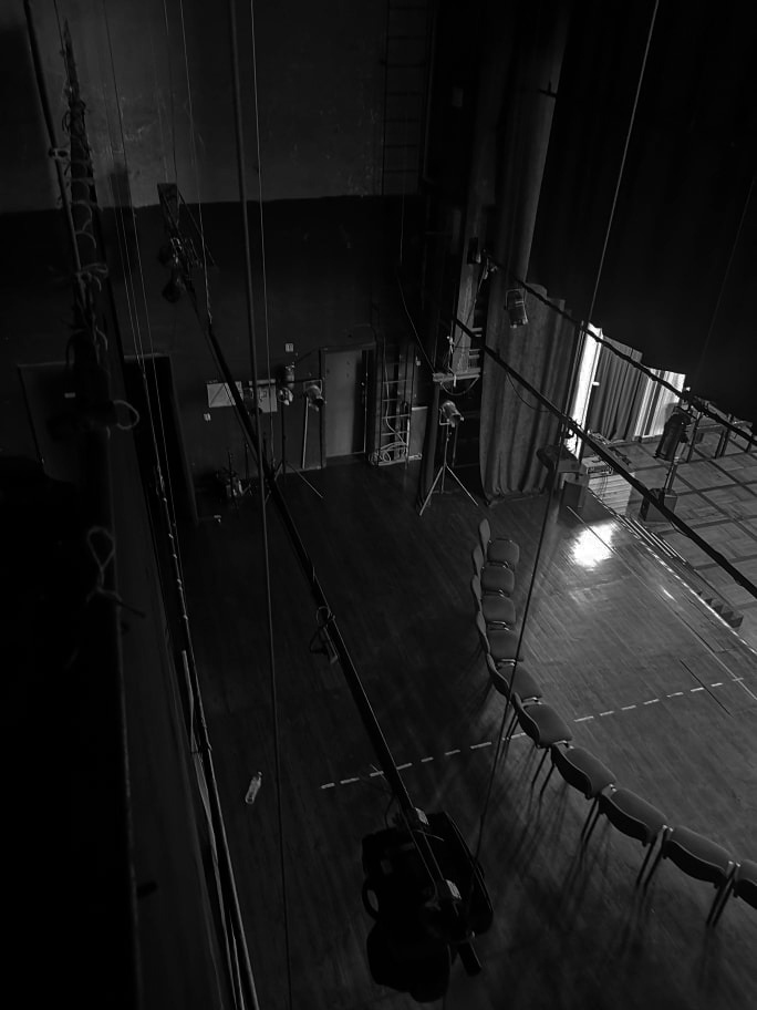 Czarno-białe zdjęcie z góry przedstawiające wnętrze teatru: drewniana podłoga z liniami, rzędy krzeseł, oświetlenie sceniczne i systemy linowe, z widocznymi elementami zaplecza.