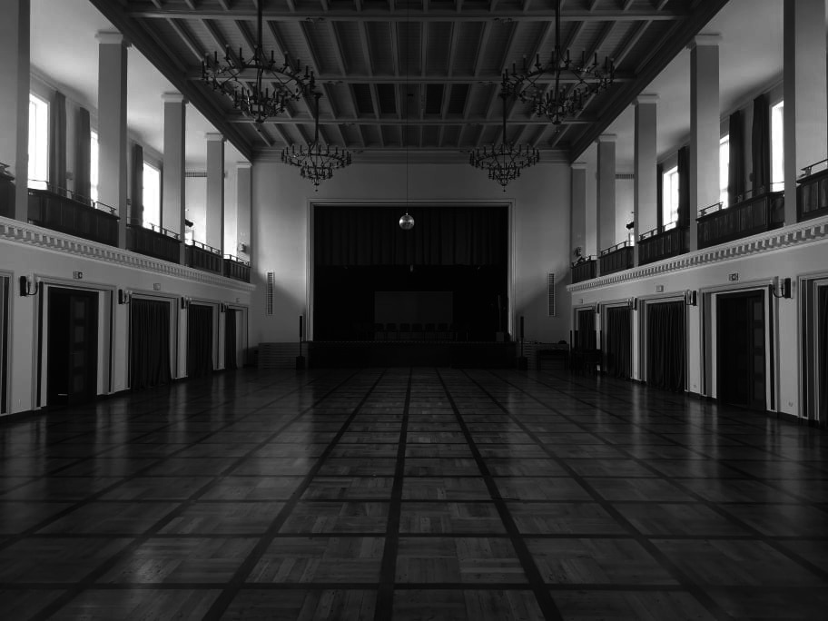 Czarno-białe zdjęcie przestronnej sali balowej z parkietem w szachownicę, kolumnami po bokach, balkonami na górze i żyrandolami zwisającymi z sufitu. Scena z ciemną kurtyną w tle.