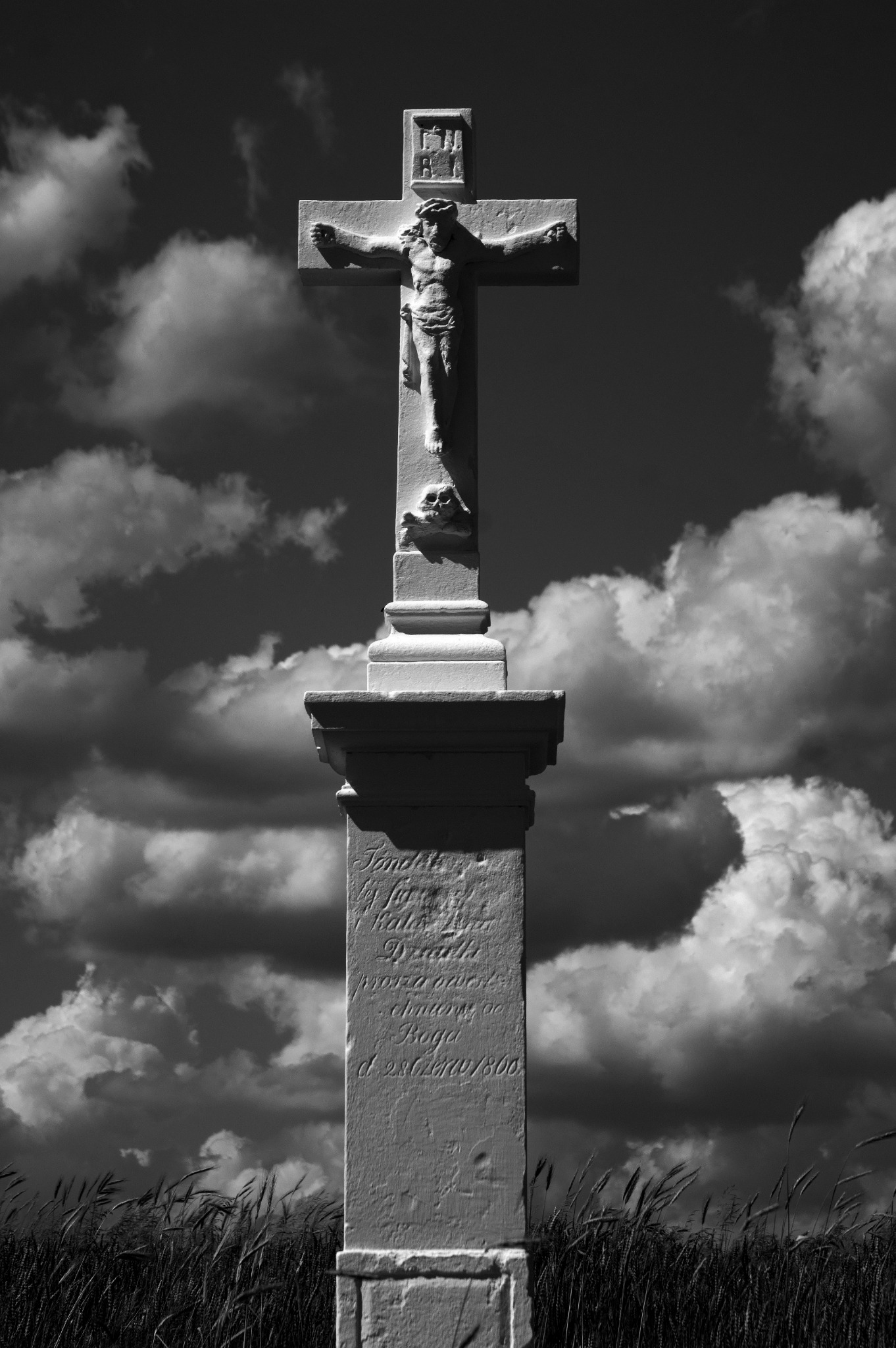 Czarno-białe zdjęcie kamiennego krzyża z Jezusem na tle pochmurnego nieba i pola pszenicy, z inskrypcjami na cokole.